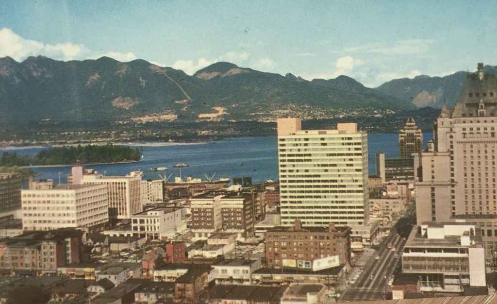 A bird's eye view of Vancouver, BC looking north from the B.C. Electric Building, showing a portion of the downtown skyline. Vancouver Harbour and the North Shore Mountains in the background. (Plastichrome, Published by Natural Colour Productions Ltd., Vancouver, BC)